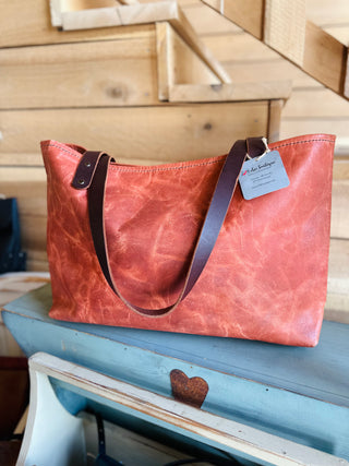 Red leather weekender tote bag with brown handles displayed on blue wooden bench in rustic setting.