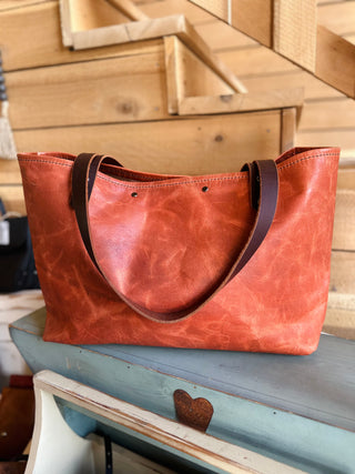 Large leather tote bag with brown handles sitting on rustic blue wooden bench in workshop setting.