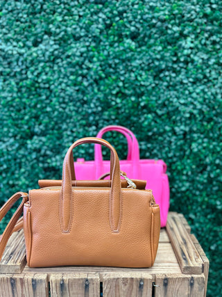 Brown leather satchel crossbody bag with compartments displayed on wooden surface with bright pink bag behind.