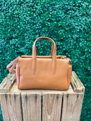 Brown leather satchel crossbody bag with dual handles displayed on rustic wooden surface against green textured background.