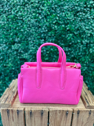 Pink leather satchel crossbody bag with handles sitting on wooden surface against green foliage background.