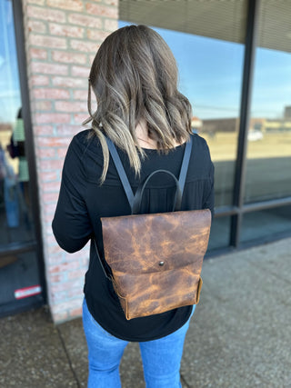 Woman wearing brown leather backpack with shoulder straps, shown from behind in casual outfit.