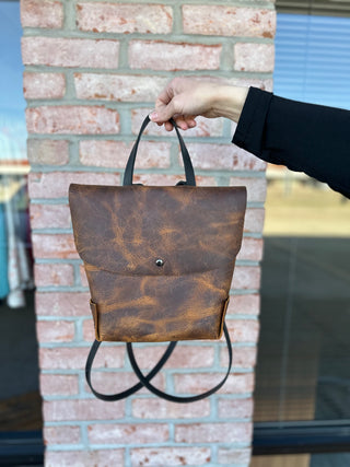 Hand holding brown leather backpack with marbled pattern and black straps against brick wall background.