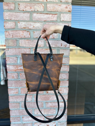 Hand holding brown leather backpack with convertible straps against brick wall background