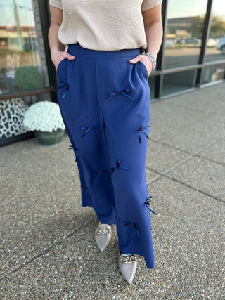Woman wearing blue wide-leg pants with decorative bow details and pockets, paired with neutral top and pointed flats.