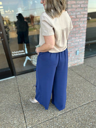 Woman wearing beige top and blue wide-leg pants standing on sidewalk outside brick building