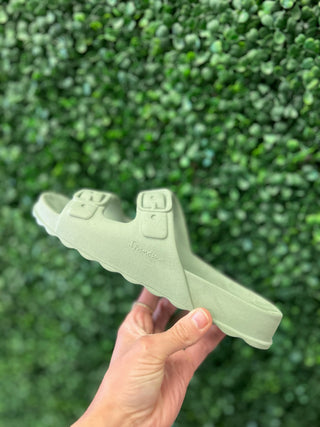 Hand holding mint green Ipanema Basic Duo sandal against blurred leafy background.
