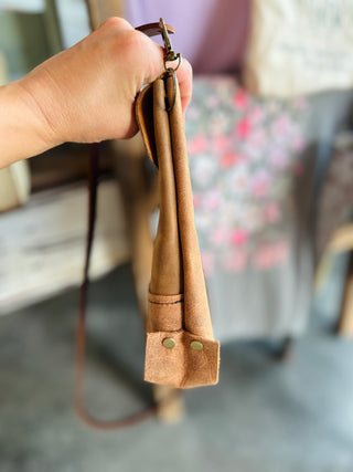 Hand holding brown leather crossbody bag with adjustable strap and metal hardware against blurred background.