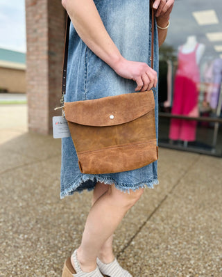 Woman wearing denim dress carrying brown leather crossbody bag with envelope flap closure and adjustable strap.