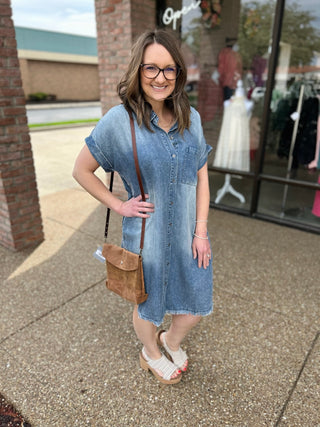 Woman wearing denim dress and glasses carrying brown crossbody bag outside boutique storefront