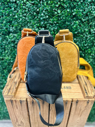 Three leather sling bags in different colors displayed on wooden crate against green foliage background.