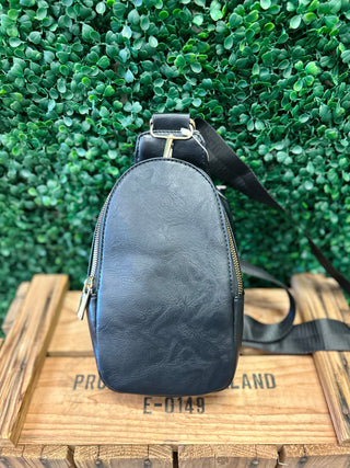 Black leather sling bag with zipper and adjustable strap displayed on wooden crate with green foliage background.