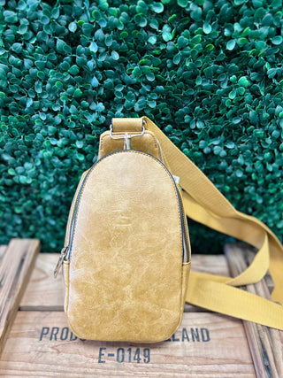 Yellow leather sling bag with crossbody strap on wooden surface against green foliage background