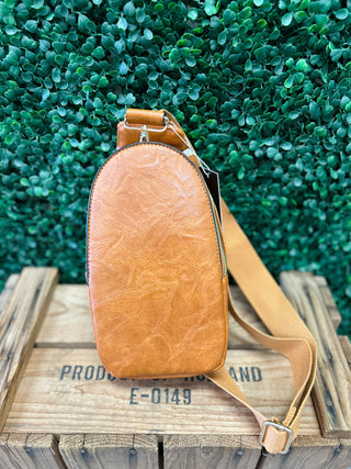 Brown leather sling bag with adjustable strap displayed on wooden crate against green foliage background.