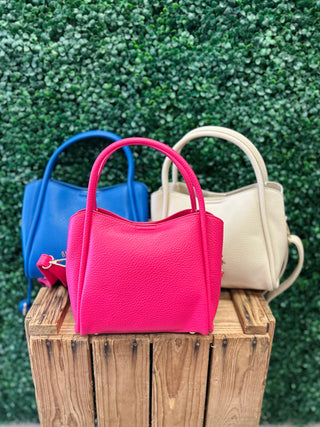 Three leather satchel handbags with top handles displayed on wooden surface against green hedge background.