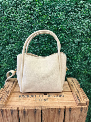Beige leather satchel handbag with dual handles and crossbody strap on wooden crate against green hedge backdrop.