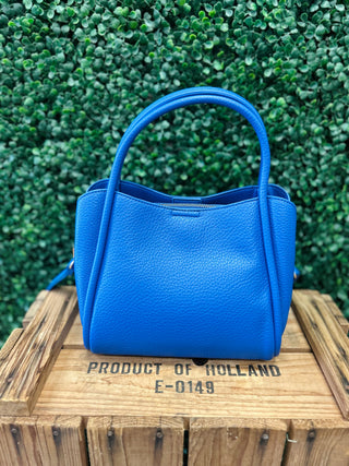 Blue leather satchel handbag with handles displayed on wooden crate against green foliage background.