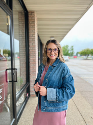 Woman wearing denim cargo snap jacket over pink dress, standing on covered walkway outside storefront.