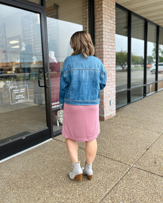 Woman wearing denim jacket and pink skirt standing outside storefront, shown from behind