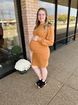 Pregnant woman wearing caramel knit maternity dress, standing outdoors by brick building with fall mums.
