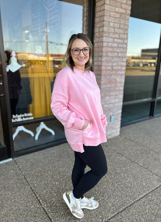 Woman wearing pink oversized pullover sweatshirt with black leggings and sneakers, standing outside storefront.