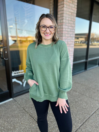 Woman wearing green oversized pullover sweater with pockets, standing outdoors near storefront windows.