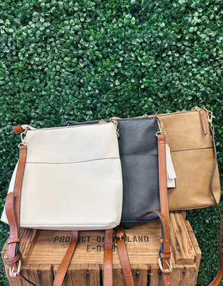 Three crossbody bags in different colors displayed on wooden crate against green foliage background