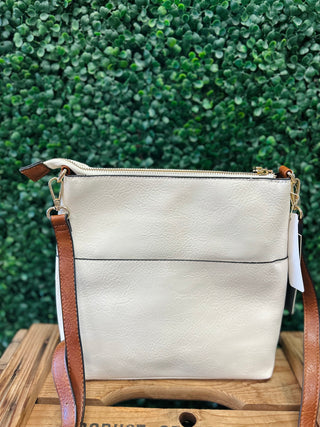 White crossbody bag with brown leather strap and gold hardware displayed on wooden surface against green foliage.