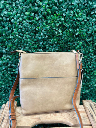 Beige crossbody bag with brown leather strap sitting on wooden surface against green foliage background.
