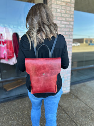 Woman wearing red leather backpack with black straps, shown from behind in casual outfit.