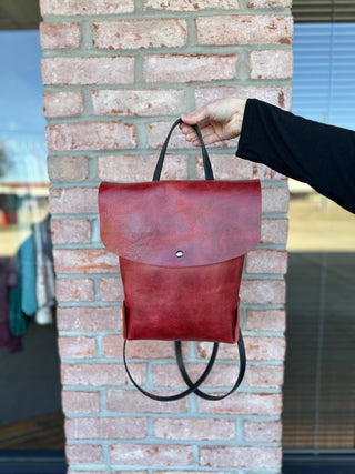 Hand holding red leather backpack with flap closure and adjustable straps against brick wall background.