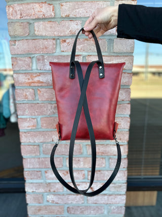 Hand holding red leather backpack with black straps against brick wall background