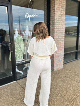 Woman wearing wide-leg textured pants with elastic waistband, paired with matching top, standing outside boutique.