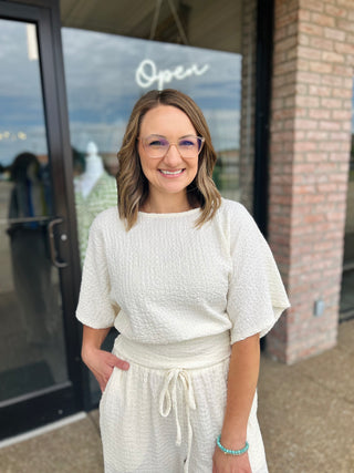 Woman wearing a textured cream short-sleeve top with relaxed fit, standing outside a boutique.