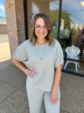 Woman wearing gray ribbed short-sleeve top and matching pants, standing outdoors with hand on hip, smiling.