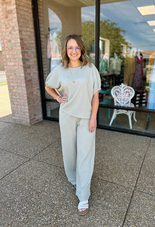 Woman wearing sage green knit top and wide-leg pants set, standing outside storefront with hands on hips.