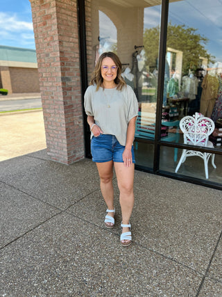 Woman wearing casual sage green top with denim shorts standing outside boutique storefront