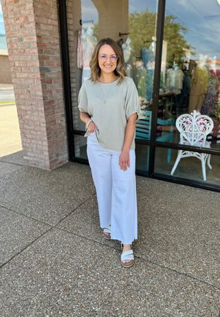Woman wearing sage green gathered short-sleeve top with white pants, standing outside boutique storefront.