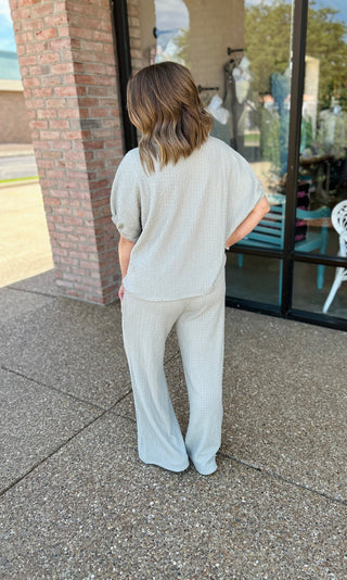 Woman wearing light colored knit top and matching pants, standing outside storefront with brick columns.