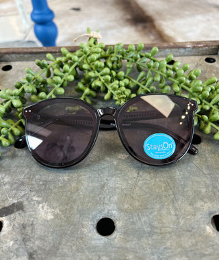Black sunglasses with dark lenses and blue brand sticker displayed on weathered wood surface with green plant.