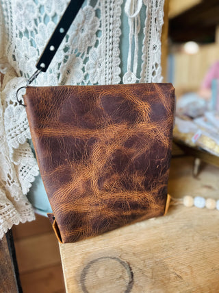 Brown leather crossbody purse with wrist strap displayed on wooden surface with lace backdrop.