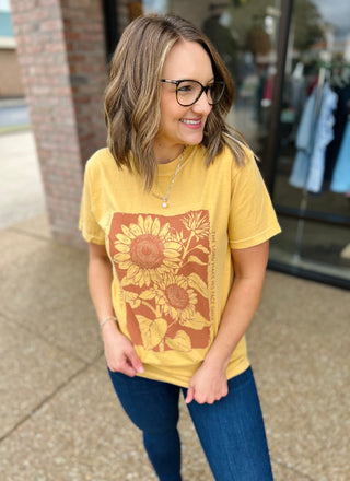 Woman wearing yellow sunflower graphic tee with jeans and glasses, smiling outdoors near storefront