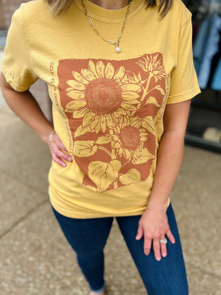 Woman wearing yellow sunflower graphic tee with botanical design and inspirational text, paired with jeans and layered nec...