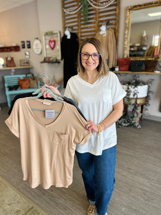 Woman in boutique holding neutral-colored v-neck pocket tees on hangers, smiling at camera