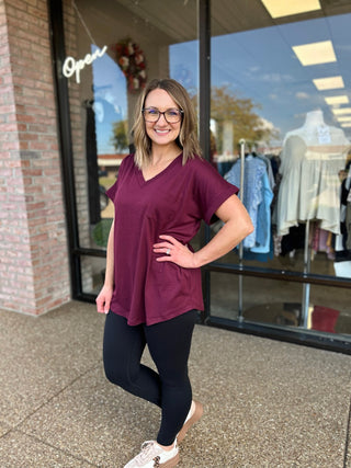 Woman wearing burgundy v-neck pocket tee and black leggings, standing outside clothing boutique storefront.