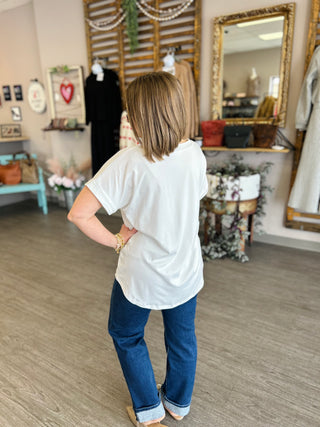 Woman wearing white pocket tee and jeans in boutique setting, shown from behind with hand on hip.