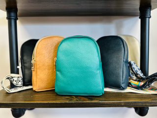 Collection of leather sling bags in various colors displayed on wooden shelf with metal frame.