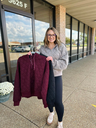 Woman wearing gray quilted quarter zip pullover holding burgundy and black versions outside storefront