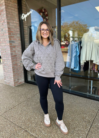 Woman wearing gray quilted quarter-zip pullover with black leggings standing outside clothing boutique storefront.