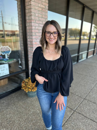 Woman wearing black long-sleeve blouse with tie detail and jeans, standing outside storefront.